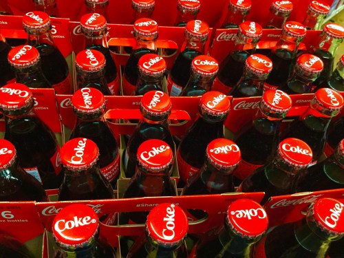 A close-up of multiple Coca-Cola bottles with red caps and packaging, arranged in a grid pattern.