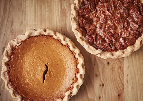 Two freshly baked pies on a wooden surface: one pumpkin pie and one pecan pie, both in golden crusts.