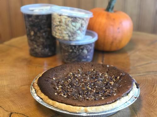 A chocolate pie topped with nuts sits on a wooden table, with jars of granola and a pumpkin in the background.