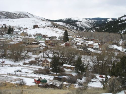 A snowy mountain town with historic buildings, trees, and hills in the background under a cloudy sky.
