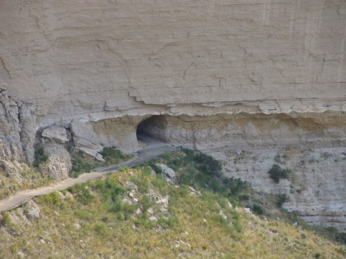 A rocky cliff with a natural cave entrance and a winding path leading into the cave, surrounded by greenery.