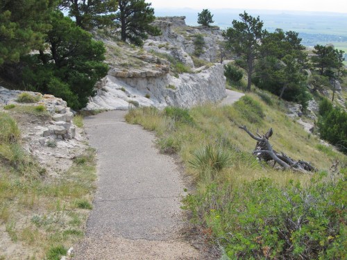 A winding path through grassy terrain, flanked by rocky cliffs and trees under a clear sky.