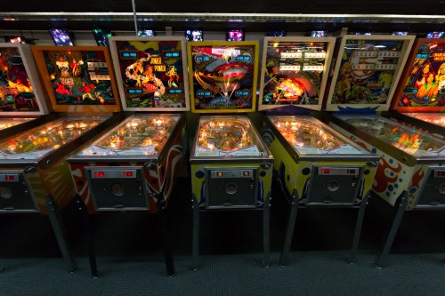 A row of colorful vintage pinball machines with illuminated playfields and vibrant artwork in a dimly lit arcade.