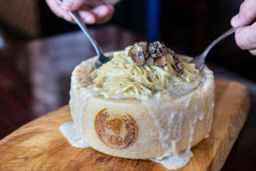 A creamy pasta dish served in a cheese wheel, topped with truffles, on a wooden board. Two forks are ready to serve.