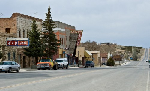A quiet street scene featuring rustic buildings, signs for a lodge and bar, and parked cars under a cloudy sky.