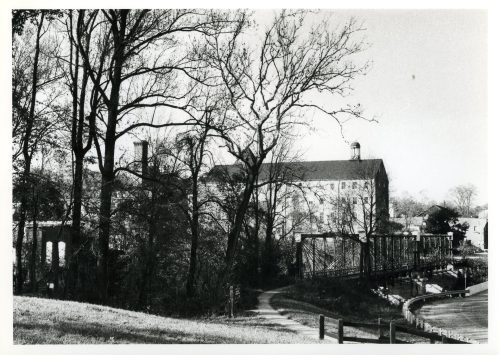 Black and white photo of a building behind trees, with a bridge and path in the foreground.