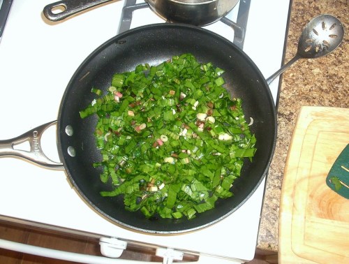 Chopped green onions sautéing in a black skillet on a stovetop, with a wooden cutting board nearby.