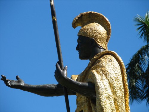 A golden statue of a warrior in profile, wearing a helmet and cape, holding a spear against a clear blue sky.