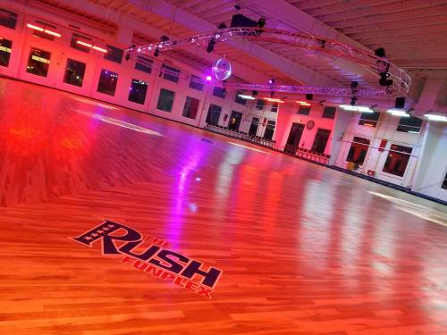 A brightly lit roller skating rink with colorful lights and a disco ball, featuring a wooden floor and "The Rush Funplex" logo.