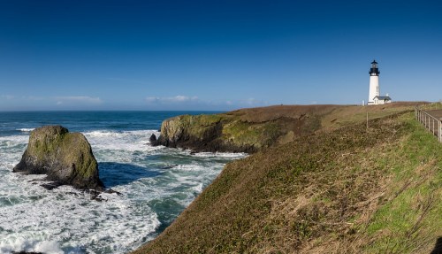 A panoramic view of a lighthouse on a cliff overlooking the ocean, with waves crashing against rocky shores under a clear blue sky.