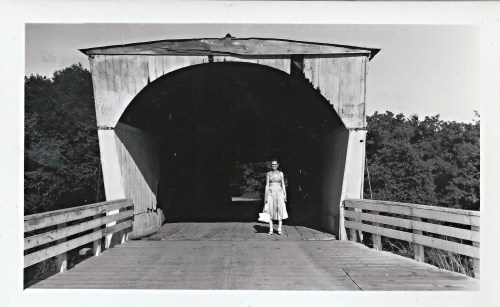 A woman in a dress stands at the entrance of a covered bridge, surrounded by trees in a black and white photo.