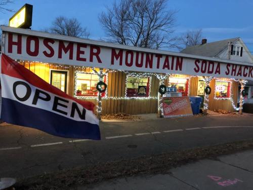 A cozy soda shack with festive lights and an "OPEN" flag, set against a winter evening backdrop.