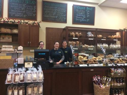 Three smiling employees stand behind a bakery counter filled with various breads and pastries, with chalkboard menus above.