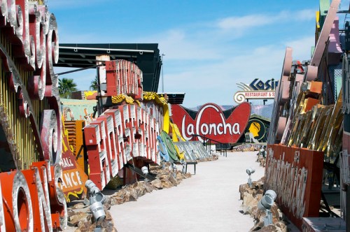 A vibrant outdoor display of vintage neon signs in a museum-like setting, with a clear blue sky above.