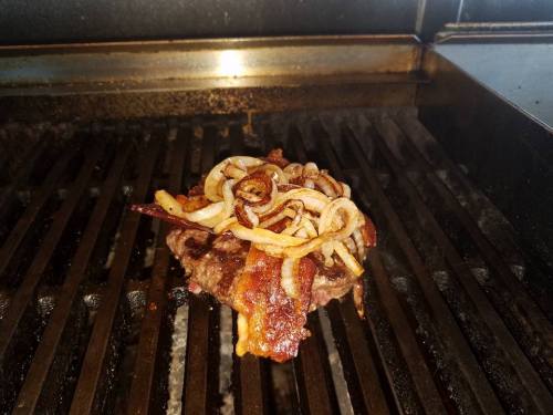 A juicy burger patty topped with crispy bacon and sautéed onions on a grill.