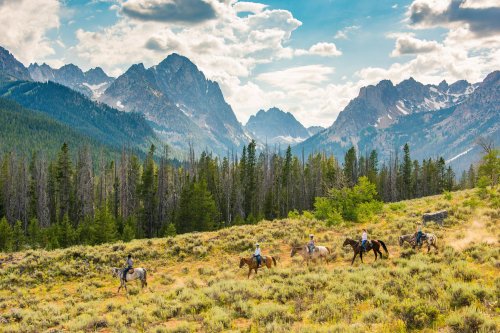 A group of horseback riders traverses a lush, green valley with majestic mountains in the background under a blue sky.