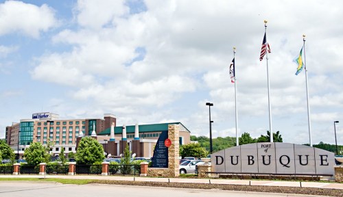 Sign for the Port of Dubuque with flags and a hotel in the background under a partly cloudy sky.