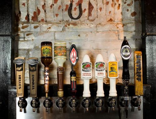 A row of various beer taps with unique designs against a rustic brick wall background.