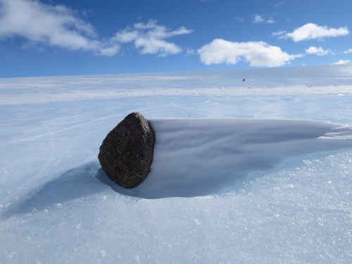 A smooth rock partially embedded in ice, with snow drifting around it under a bright blue sky and fluffy clouds.