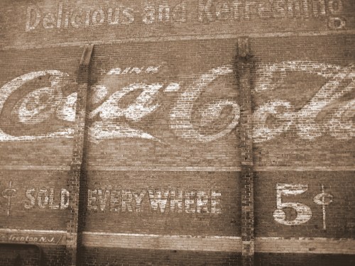 Faded vintage Coca-Cola advertisement on a brick wall, featuring the slogan "Delicious and Refreshing" and pricing.