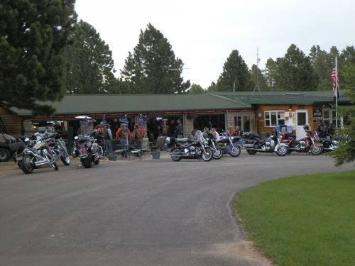 A group of motorcycles parked outside a rustic lodge surrounded by trees, with people gathered on the porch.