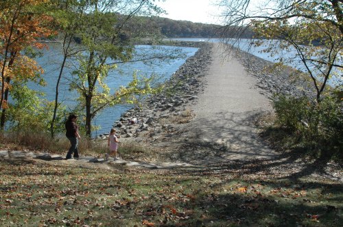 A woman and a child walk along a path by a lake, with a rocky dam and autumn foliage in the background.