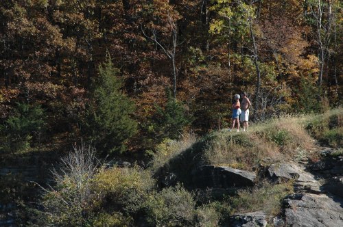 Two people stand on a rocky ledge surrounded by colorful autumn trees.