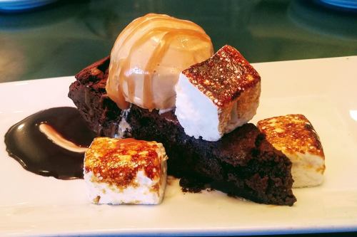 A slice of rich brownie topped with ice cream, caramel drizzle, and toasted marshmallows on a white plate.