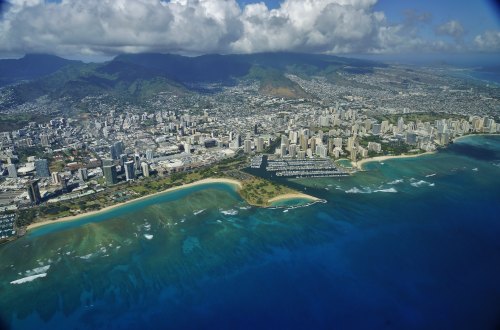 Aerial view of a coastal city with skyscrapers, beaches, and mountains under a partly cloudy sky.