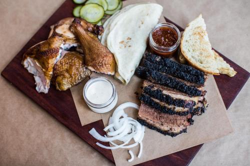 A wooden platter featuring smoked brisket, chicken, pickles, bread, and sauces, garnished with sliced onions.