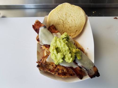 A sandwich with grilled chicken, melted cheese, and guacamole on a soft bun, served in a paper tray.