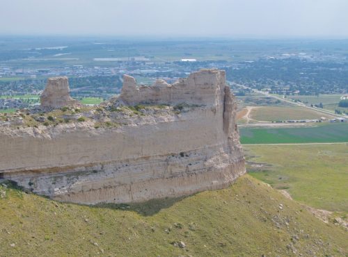 A panoramic view of a rocky cliff with flatlands and a distant town visible in the background under a hazy sky.