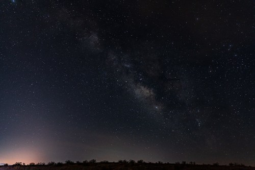 A clear night sky filled with stars and the Milky Way, with a faint glow on the horizon.