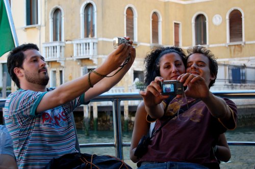 Three friends take photos of each other on a boat in Venice, with historic buildings in the background.