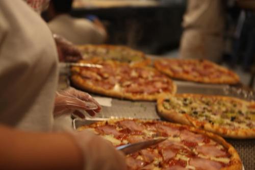 A person in gloves slices freshly baked pizzas on a countertop, with various toppings visible.