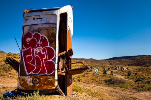 A rusted, overturned vehicle covered in graffiti, set against a desert landscape with distant hills.