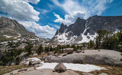 A scenic mountain landscape featuring rocky peaks, scattered clouds, and patches of snow among green trees.