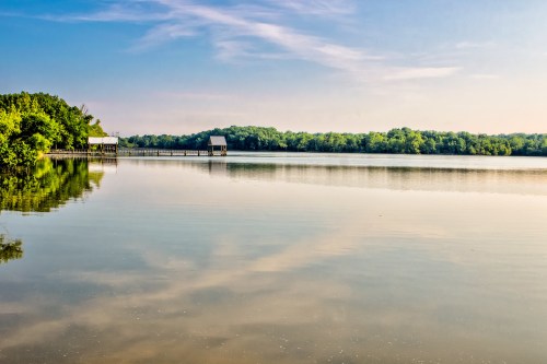 A serene lake scene with calm waters reflecting trees and a small dock under a clear blue sky.