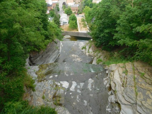 Aerial view of a waterfall cascading over rocky terrain, surrounded by lush green trees and a glimpse of a town below.