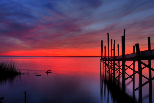 A serene sunset over calm waters, with a silhouetted wooden pier and colorful clouds reflecting in the water.