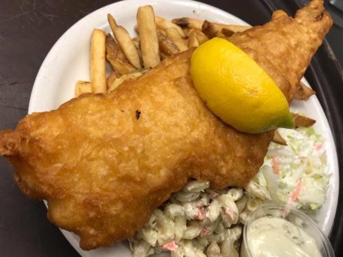 A plate of golden-brown fried fish with a lemon slice, served with fries, coleslaw, and a side of creamy dressing.