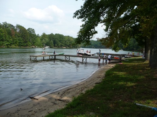 A serene lake scene with people on a dock, boats nearby, and lush greenery surrounding the water.