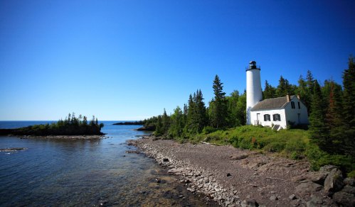 A white lighthouse stands on a rocky shore, surrounded by green trees and a clear blue sky over the calm water.