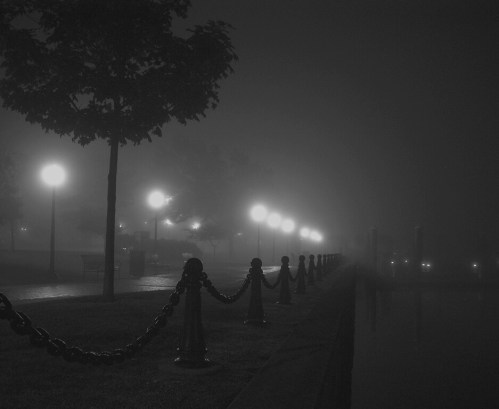 A foggy night scene with street lamps illuminating a path beside a calm waterway, surrounded by trees and chains.