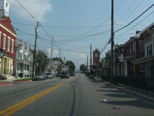 A quiet street lined with historic buildings, power lines overhead, and a clock tower in the distance under a blue sky.