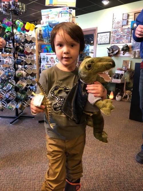 A young child holds a plush dinosaur and a small toy, smiling in a gift shop filled with various items.