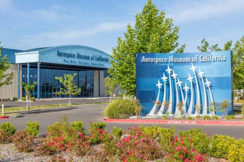 Exterior view of the Aerospace Museum of California, featuring a sign and landscaped area with trees and flowers.