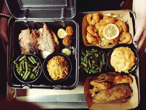 Two takeout containers filled with barbecue brisket, fried shrimp, sides of green beans, macaroni, and fried fish.