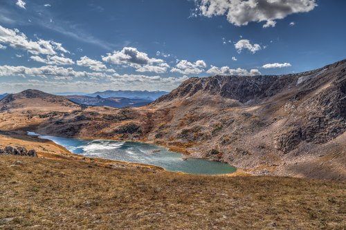 A scenic view of a mountain landscape featuring a serene lake surrounded by rocky terrain and distant peaks under a blue sky.