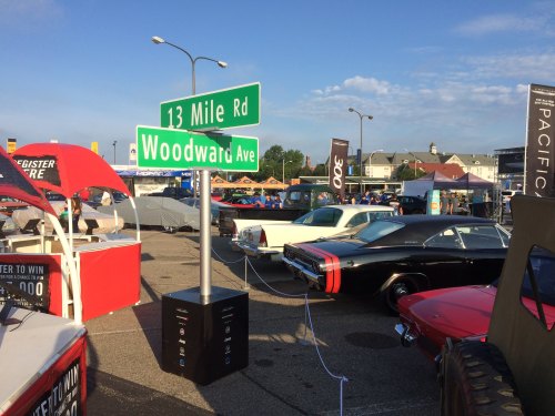 Street sign at a car show, with classic cars and tents in the background under a clear blue sky.